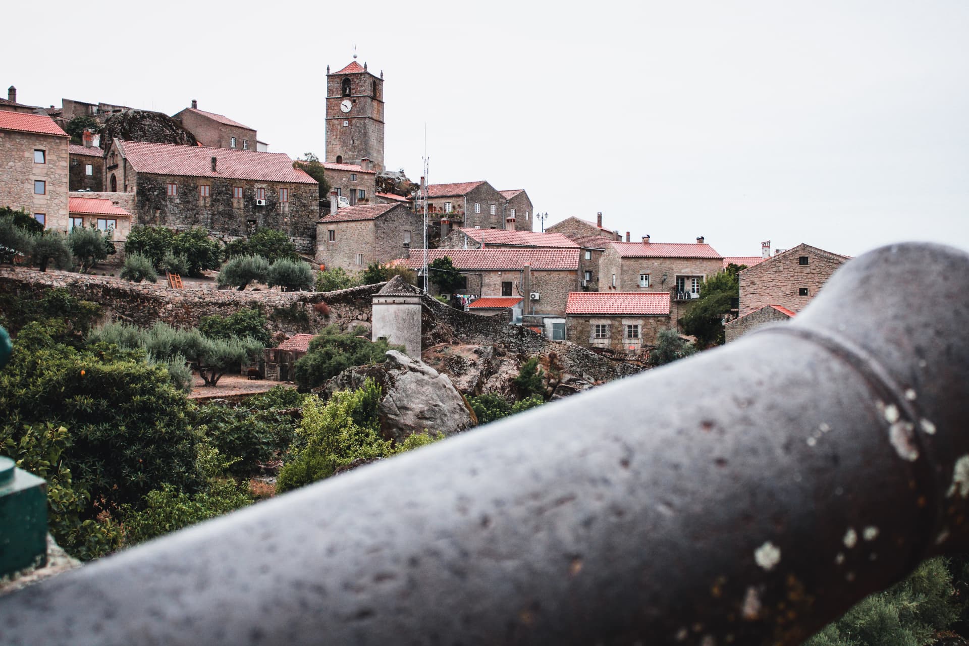 The village of Monsanto on its granite hill, with the clock tower of the old castle above the rooftops, seen past an old cannon, photographed by Ricardo Tito